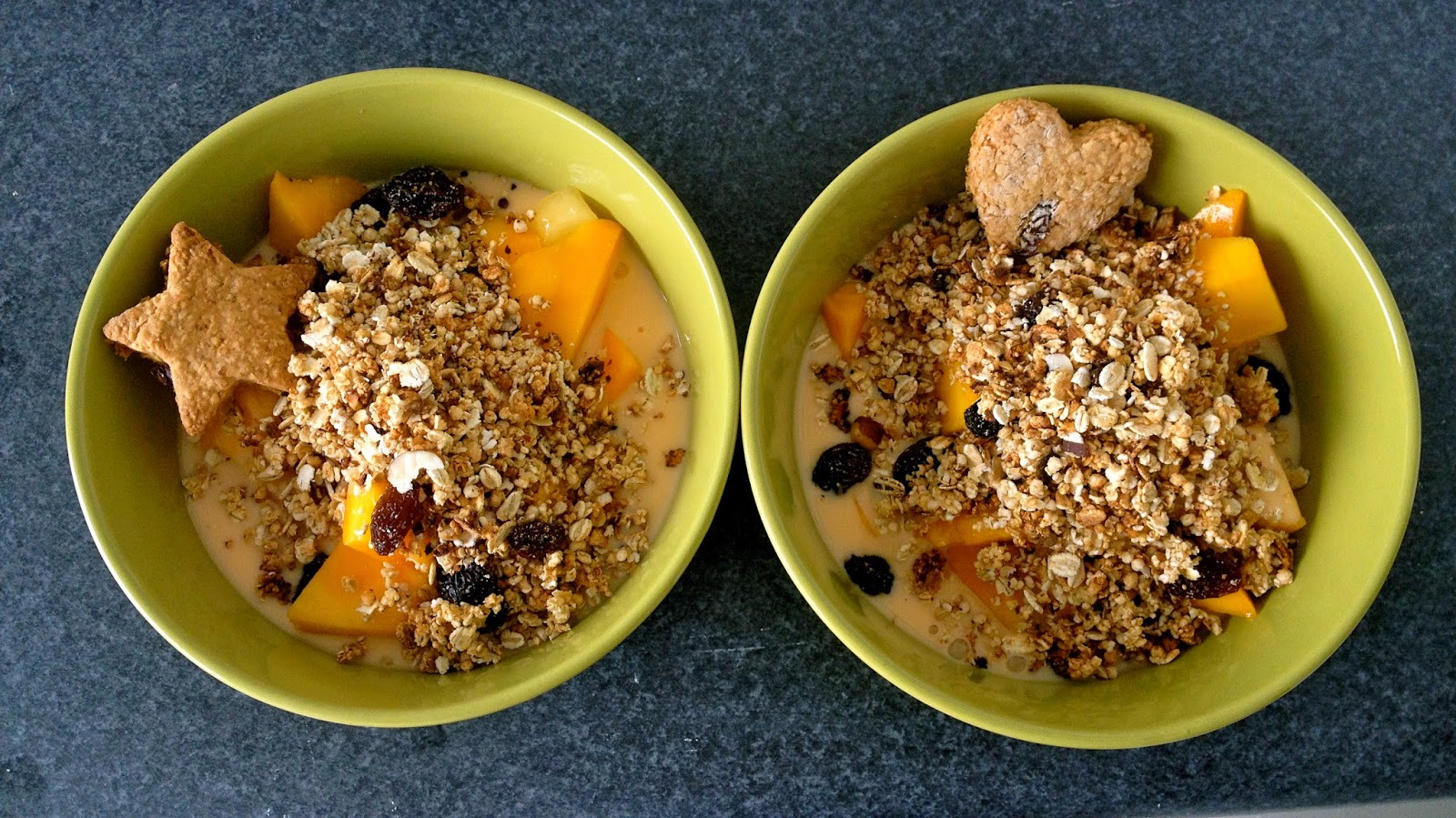 Two green bowls of granola with mango pieces, dried fruit, and cookie toppings shaped like a star and heart, on a dark countertop